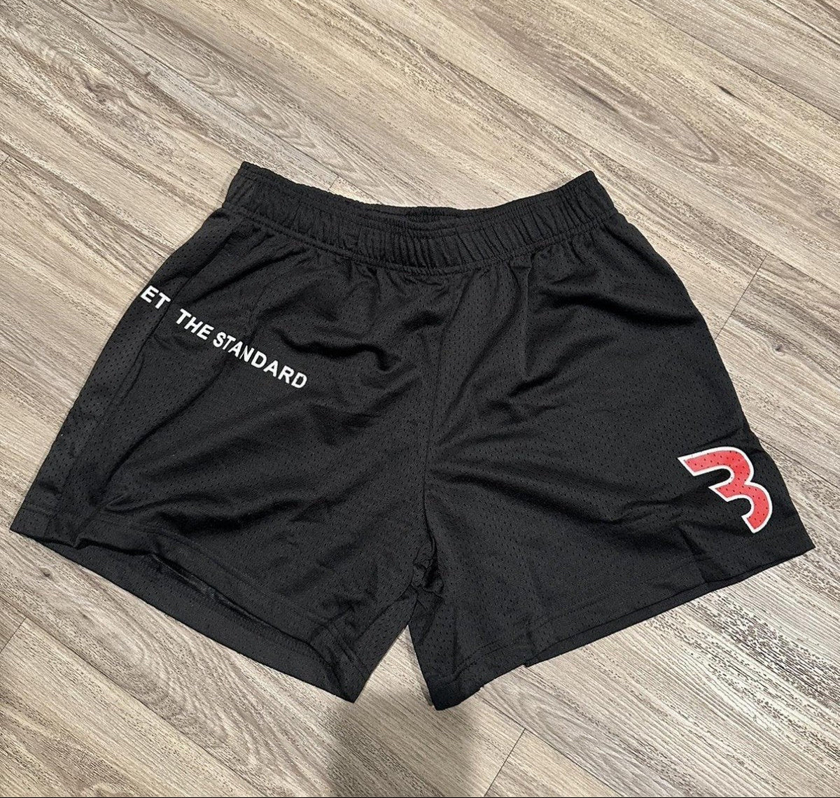 mesh shorts de la merch de CBUM en color negro, con una B de su logo en blanco y rojo y la frase "set the standard" en blanco
